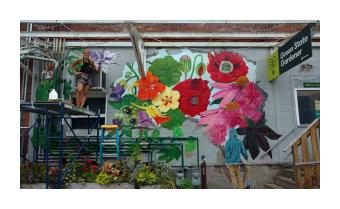 a mural of lush hyper realistic flowers in reds, yellows, pinks, and greens on the side of a grey brick building 