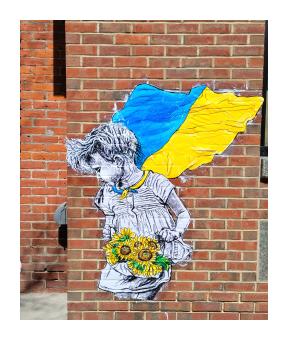 a mural of a little girl in black and white with a cape made from the blue and yellow Ukrainian flag holding a bouquet of yellow sunflowers 