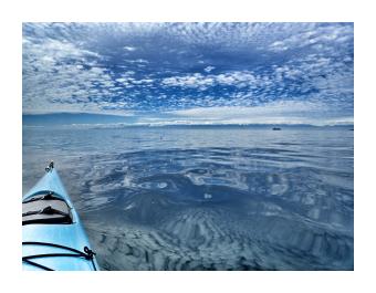 A photograph of the nose of a blue kayak on a body of water that reflects the cloud strewn sky above