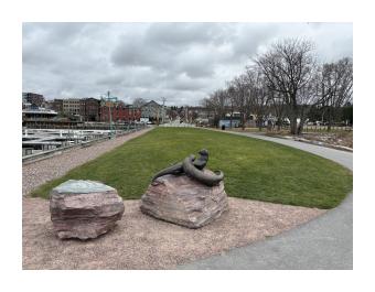 A picture of a park with two large boulders centered. On one of the boulders there are two large otter sculptures.