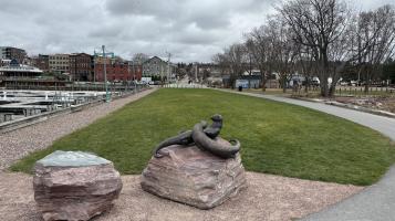 A picture of a park with two large boulders centered. On one of the boulders there are two large otter sculptures.